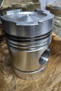 Daihatsu DL-20 Engine Piston Profile Side perspective of a polished metallic piston showing the piston ring grooves and wrist pin hole, resting on crumpled brown paper packaging.