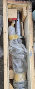 Engine Camshaft in Wooden Crate Top-down view of a long metallic engine camshaft partially covered in protective plastic, secured inside a wooden shipping crate.
